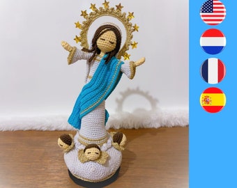 May include: A crocheted figurine of a figure with brown hair, wearing a white robe and a blue sash, featuring a gold halo adorned with stars. The base is a white cloud shape decorated with three small cherub faces with brown hair and gold wings.