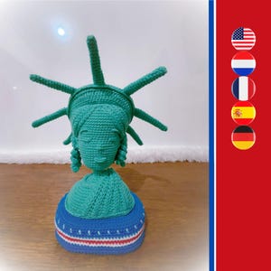 Può includere: Una statuetta all'uncinetto color verde acqua della Statua della Libertà con una corona a sette punte, un corpetto drappeggiato e una base a strisce orizzontali blu, bianche e rosse. A destra, una barra rossa verticale mostra cinque bandiere.