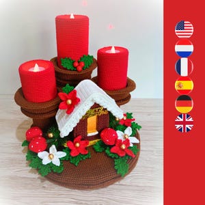 May include: A crocheted Christmas centrepiece with three red candles with faux flames, a miniature house, and festive decorations. The piece is adorned with red berries, white flowers, and green leaves, creating a cosy holiday atmosphere.