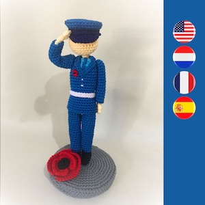 Könnte beinhalten: Eine gehäkelte Puppe in blauer Uniform und einer dunkelblauen Mütze, die mit einer roten Mohnblume zu ihren Füßen salutiert.
