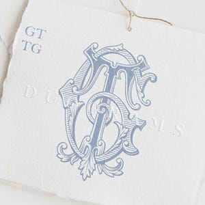 May include: A square piece of textured white paper featuring a blue ornate monogram of the letters GT and TG. The paper has a deckled edge and a thin brown string attached at the top. The text GT, TG, and DUOGRAMS are visible on the paper.