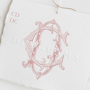 May include: A pink monogram with the letters "CD" intertwined in a decorative, ornate font. The letters are surrounded by floral designs. The text "DUOGRAMS" is printed below the monogram.