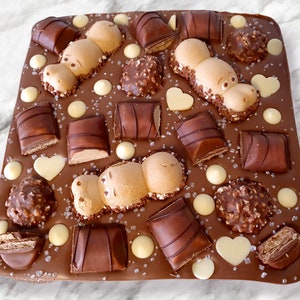 May include: A rectangular chocolate bark with various chocolate candies and decorations. The bark is adorned with Kinder Bueno bars, Ferrero Rocher, white chocolate hearts, and round white chocolate pieces. Two hippo-shaped candies are also present, sprinkled with sugar crystals.