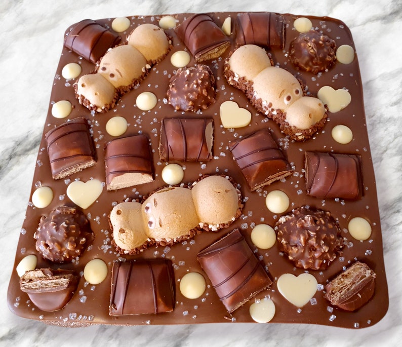 May include: A rectangular chocolate bark with various chocolate candies and decorations. The bark is adorned with Kinder Bueno bars, Ferrero Rocher, white chocolate hearts, and round white chocolate pieces. Two hippo-shaped candies are also present, sprinkled with sugar crystals.