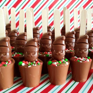 May include: A selection of chocolate Santa Claus figures, each with a wooden stick, presented on a red and white striped surface. The Santas sit atop chocolate cups with sprinkles. A festive treat for Christmas.
