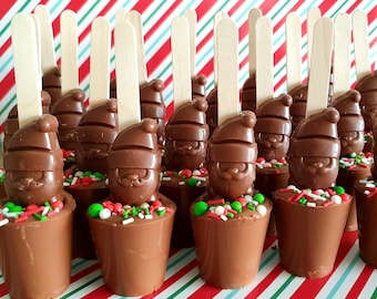 May include: A collection of chocolate Santa Claus figures on a red and white striped background. Each Santa is atop a chocolate cup filled with sprinkles, and has a wooden stick. Festive treats for the holiday season.