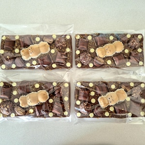 May include: Four chocolate bars in clear packaging. Each bar is a mix of dark chocolate, white chocolate chips, and various chocolate pieces. A light brown hippo-shaped candy sits on top of each bar.