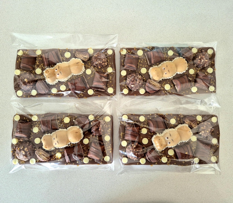 May include: Four chocolate bars in clear packaging. Each bar is a mix of dark chocolate, white chocolate chips, and various chocolate pieces. A light brown hippo-shaped candy sits on top of each bar.