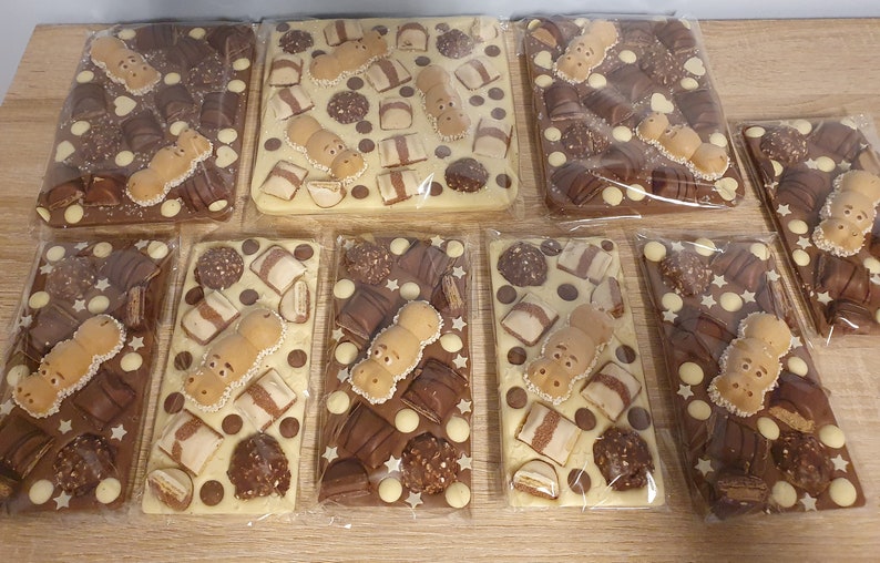 May include: Several chocolate bars, each wrapped in clear plastic. The bars are rectangular and topped with various chocolates, including hippo-shaped candies, chocolate pieces, and white chocolate stars. The chocolate bars are on a wooden surface.