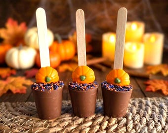 May include: Three chocolate cups on wooden sticks, each topped with a small orange pumpkin candy and a layer of purple, orange, and black sprinkles. The cups are arranged in a row, featuring a smooth brown chocolate exterior and a flat top.