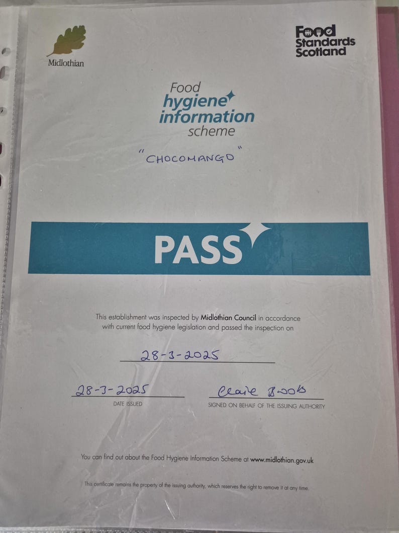 May include: A food hygiene information scheme certificate from Midlothian, Scotland, with the text "CHOCOMANGO" and the word "PASS" in large letters. The certificate is dated March 28, 2025, and signed by Clare Boos.