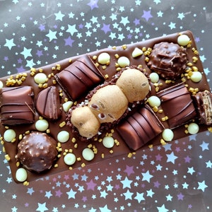 May include: A rectangular chocolate bar topped with various chocolates, including truffles, wafers, and candies. The bar is decorated with white chocolate drops and gold sprinkles. The background is a dark surface with a pattern of white and purple stars.