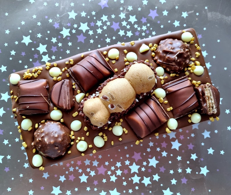 May include: A rectangular chocolate bar topped with various chocolates, including truffles, wafers, and candies. The bar is decorated with white chocolate drops and gold sprinkles. The background is a dark surface with a pattern of white and purple stars.