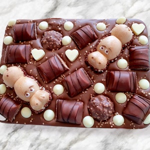 May include: A rectangular chocolate bar with various toppings. The bar is covered in dark chocolate and decorated with chocolate candies, white chocolate pearls, heart-shaped candies, and two hippo-shaped meringues.