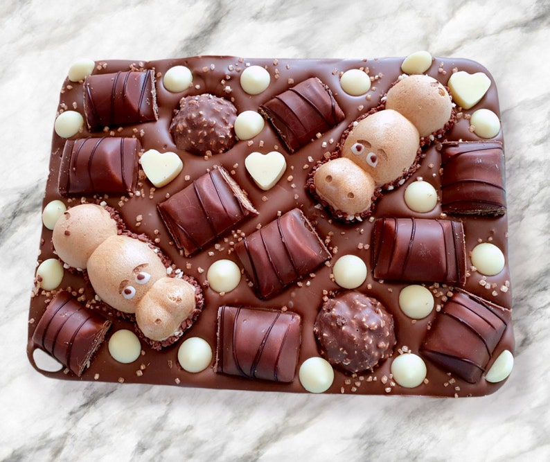 May include: A rectangular chocolate bar with various toppings. The bar is covered in dark chocolate and decorated with chocolate candies, white chocolate pearls, heart-shaped candies, and two hippo-shaped meringues.