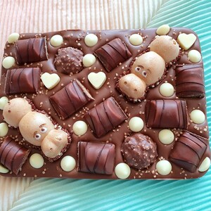May include: A rectangular chocolate bar with various toppings. The bar is decorated with chocolate candies, white chocolate drops, heart-shaped chocolates, and two hippo-shaped chocolates. The background is a light pink and teal fabric.