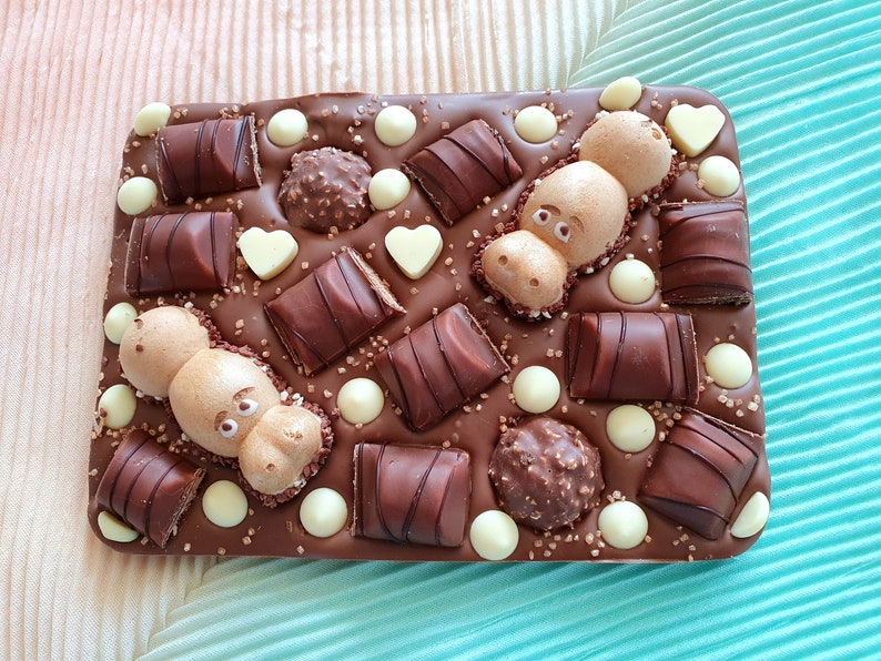 May include: A rectangular chocolate bar with various toppings. The bar is decorated with chocolate candies, white chocolate drops, heart-shaped chocolates, and two hippo-shaped chocolates. The background is a light pink and teal fabric.