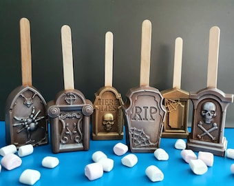 May include: Six chocolate tombstone-shaped pops on wooden sticks, featuring metallic bronze and gold finishes with molded details including a spider, a skull, crossbones, spiderwebs, and the text "HERE LIES" and "RIP," surrounded by white marshmallows.