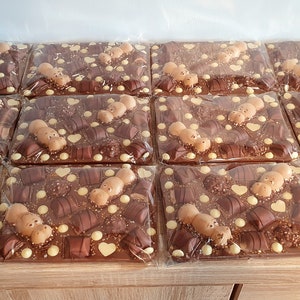 May include: A collection of individually wrapped chocolate bars, each featuring a variety of chocolates, white chocolate drops, and heart-shaped decorations. The bars are arranged on a wooden surface.