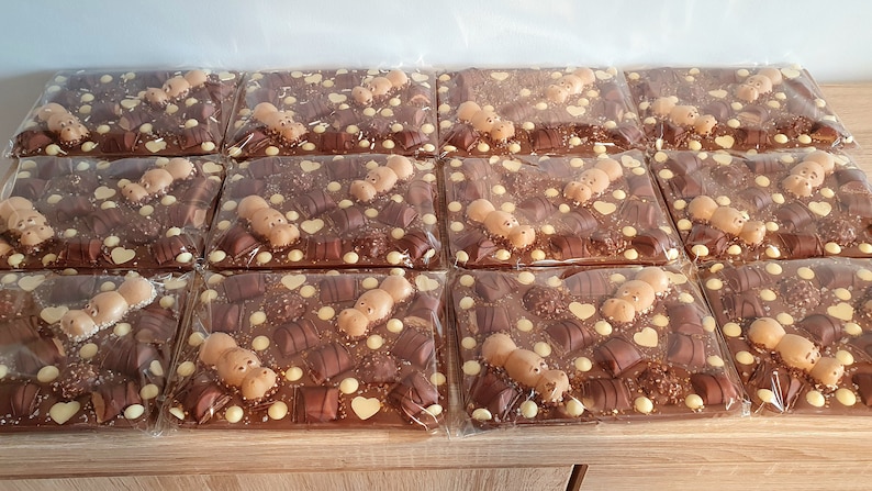 May include: A collection of individually wrapped chocolate bars, each featuring a variety of chocolates, white chocolate drops, and heart-shaped decorations. The bars are arranged on a wooden surface.