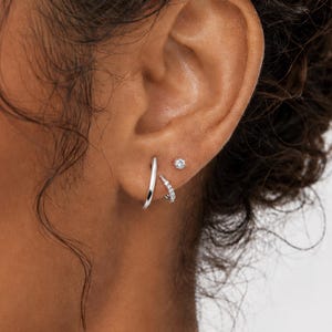 May include: Close-up of an ear adorned with three silver earrings. One is a small stud with a single clear stone, and the other two are hoop and curved designs. The earrings are set against the skin.