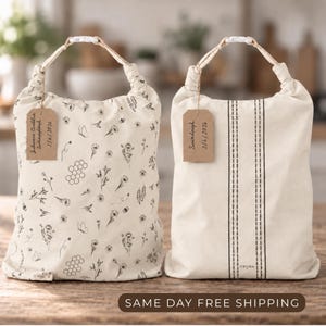May include: Two off-white fabric storage bags with drawstring closures and white plastic toggles. The left bag features a black botanical and honeycomb print. The right bag has two vertical black dashed lines. Each bag includes a brown paper tag.