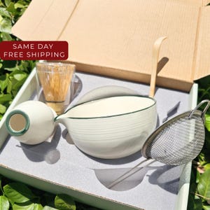 May include: A matcha tea set in a cardboard box containing a white ceramic bowl with a green rim, a white ceramic pitcher, a bamboo whisk in a clear glass holder, a bamboo scoop, and a stainless steel mesh strainer with a long handle.