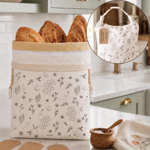 May include: A cream-colored fabric bread bag featuring a black line-art pattern of honeycombs, flowers, butterflies, and hummingbirds. The bag has a tan top band, a white zipper closure, and side drawstrings. It is filled with three loaves of bread.