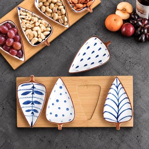 May include: A set of leaf-shaped ceramic serving dishes with blue and white patterns, including polka dots and botanical motifs, arranged on rectangular bamboo trays alongside bowls filled with nuts and fresh fruit.