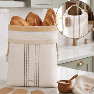 May include: A cream-colored fabric bread bag with a zipper closure, featuring two vertical black dashed stripes and the text UWOMA printed near the bottom. The bag is filled with three loaves of crusty bread and includes a hanging tag.