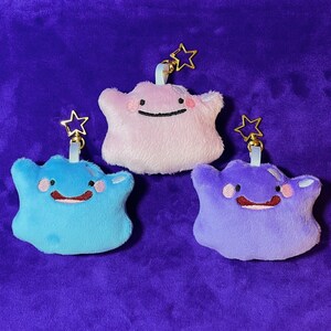 May include: Three plush Ditto keychains in light blue, pink, and purple. Each has a smiling face, rosy cheeks, and a gold star-shaped clasp. The keychains are on a purple background.