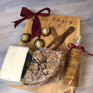 Puede incluir: Una tabla de cortar de madera con el nombre "Flia Rodriguez Acosta" grabado en ella. La tabla está decorada con un lazo rojo, trufas de chocolate, un trozo de queso, galletas y una cuchara de madera.