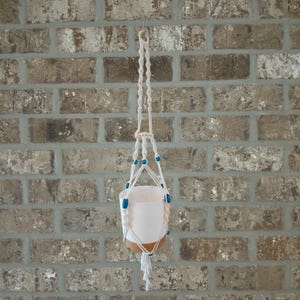 May include: A macrame plant hanger made of off-white cord featuring a series of knotted patterns and bright blue beads. The hanger supports a cylindrical pot that is white on the top half and a solid tan color on the bottom half, ending in a tassel.