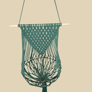 May include: A dark green macrame wall hanging featuring a triangular knotted pattern suspended from a light-colored wooden dowel. The design incorporates a circular black metal wire frame integrated into the lower section of the cord structure.
