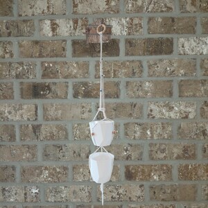 May include: A double-tiered macrame plant hanger featuring two white geometric ceramic pots suspended by off-white cotton cords. The structure includes a wooden ring at the top for hanging and small decorative beads integrated into the cord design.