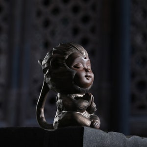 May include: A small, brown ceramic monkey figurine with a closed-eye expression, sitting in a meditative pose with hands clasped together. The monkey has a small, round, gold-colored dot on its forehead.
