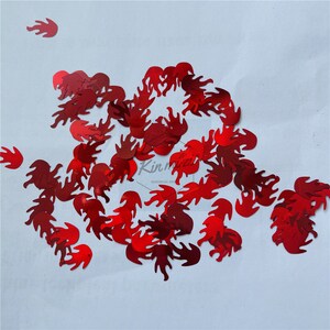 May include: Red glitter flame shaped confetti. The confetti is scattered on a white surface.