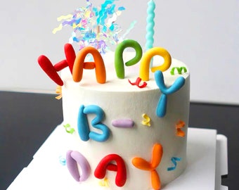 Könnte beinhalten: Ein weiß glasierter Kuchen, verziert mit bunten, abgerundeten, tonartigen Buchstaben, die HAPPY B-DAY bilden. Der Kuchen ist mit einer hellblauen Spiral-Kerze, irisierenden Lametta-Akzenten und kleinen, verstreuten, bunten Konfettistücken versehen.