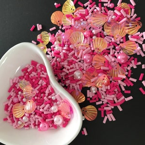 Puede incluir: Sprinkles rosas y naranjas con cuentas de perla y formas de conchas iridiscentes. Los sprinkles están en un tazón blanco en forma de corazón.