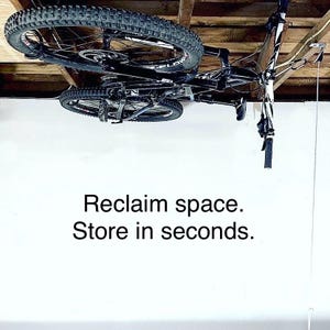 May include: A black mountain bike suspended from a wooden ceiling using a pulley storage system. The bike features knobby tires and a black frame. The text Reclaim space. Store in seconds. is displayed in black font on the white wall below.