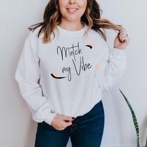 May include: White sweatshirt with the text "Match my Vibe" in black script with a black and peach abstract design.