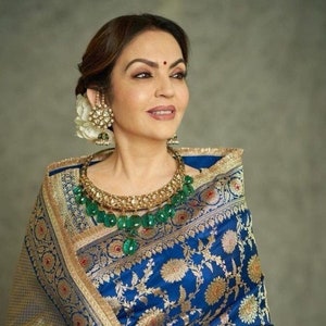 May include: A woman wearing a blue and gold patterned saree with a green gemstone necklace. She has a white flower in her hair and is wearing gold earrings.