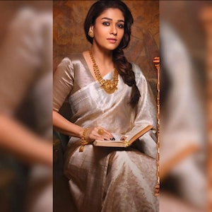 May include: A woman wearing a white and gold sari with a gold necklace and earrings. She is holding a book in her hands.