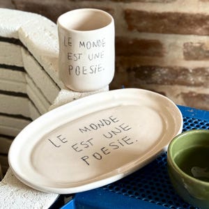 May include: A ceramic set featuring a white oval platter and a cylindrical cup, both with the handwritten phrase "LE MONDE EST UNE POESIE." A small green bowl is also visible. The items are on a blue surface.