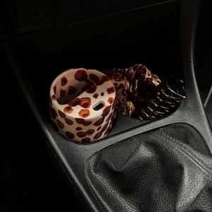 May include: A ceramic cup with a white base and brown spots, a leopard print scrunchie, and a dark brown hair clip are in a car's console. The cup has a unique design with a cutout and two holes.