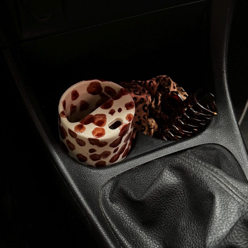 May include: A ceramic cup with a white base and brown spots, a leopard print scrunchie, and a dark brown hair clip are in a car's console. The cup has a unique design with a cutout and two holes.