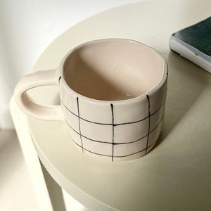 Puede incluir: Una taza de cerámica blanca con un patrón de cuadrícula negra. La taza tiene un asa redondeada y una abertura ancha.