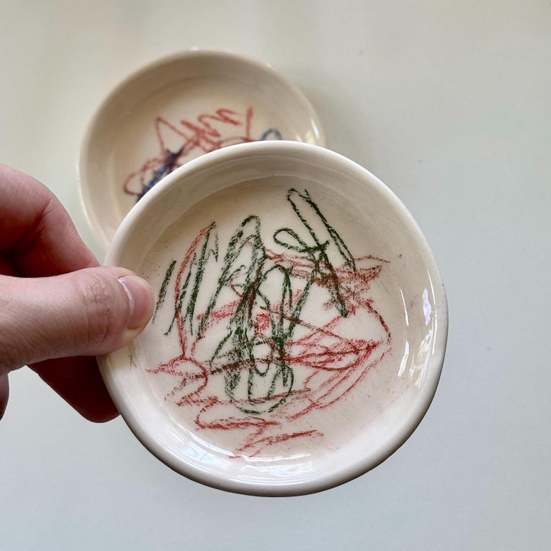 Pu&ograve; includere: Due piatti in ceramica bianca con disegni fatti a mano in rosso, verde e blu. I piatti sono piccoli e rotondi, perfetti per contenere gioielli o altri piccoli oggetti.