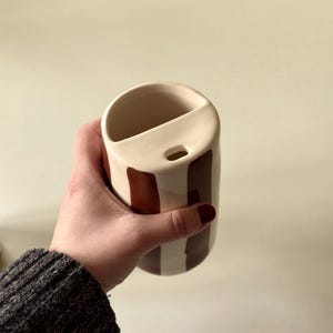 Può includere: Un vaso in ceramica color crema con strisce marroni verticali. Il vaso ha una forma unica con una parte superiore curva e un piccolo foro. Il vaso è tenuto in una mano.