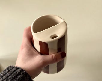Peut inclure: Un vase en céramique crème avec des rayures marron verticales. Le vase a une forme unique avec un dessus incurvé et un petit trou. Le vase est tenu dans une main.